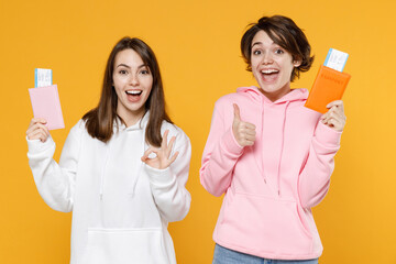 Wall Mural - Excited funny two young traveler tourist women friends 20s in casual white pink hoodies hold passport tickets boarding pass showing thumb up okay gesture isolated on yellow background studio portrait.