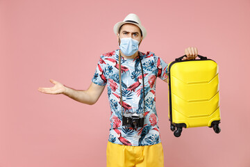 Wall Mural - Puzzled young traveler tourist man in sterile mask to safe from coronavirus hold suitcase spreading hands isolated on pink background. Passenger traveling on weekends. Air flight journey concept.