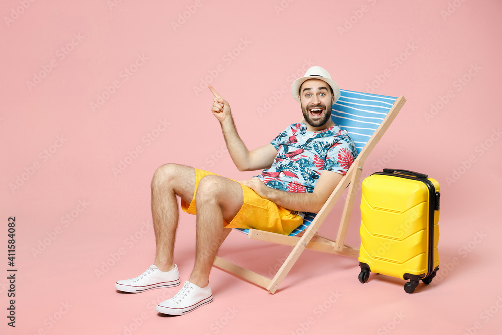 Full length of excited traveler tourist man in summer clothes hat sit on deck chair point index finger aside up isolated on pink background. Passenger traveling on weekend. Air flight journey concept. - obrazy, fototapety, plakaty 