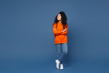 Wall Mural - Full length of attractive smiling young african american woman in casual basic bright orange sweatshirt standing holding hands crossed looking aside isolated on blue color background studio portrait.