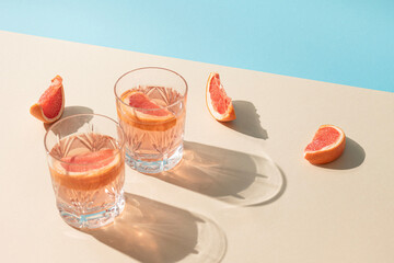 Two glasses of drink with slices of fresh grapefruit against bright beige and blue background. Creative minimal summer concept. Sunny day shadows.