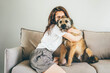 © Mariia Korneeva - Positive brunette lady in white t-shirt with glasses hugs and strokes adorable old grey dog sitting on comfortable couch in light living room at home.