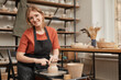 © Seventyfour - Warm toned portrait of young female potter smiling at camera while working on pottery wheel in workshop and enjoying arts and crafts, copy space