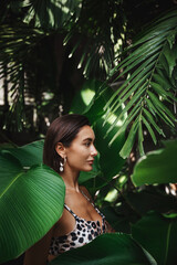 Wall Mural - Profile of tanned sexy woman in bikini, wearing earrings, standing in jungle tropical leaves and palm trees on summer vacation. Concept of tourism and tropic holidays