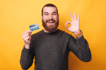 Wall Mural - Cheerful young man with beard holding credit card and showing ok gesture over yellow background.