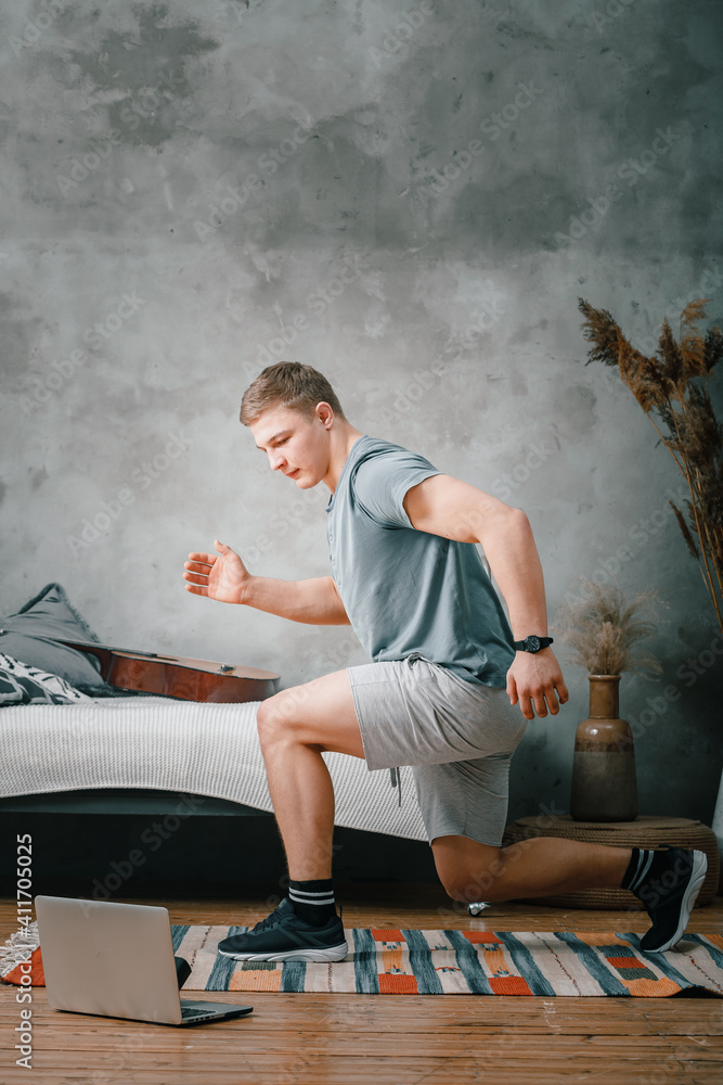 A cheerful athlete with blond hair lunges in the bedroom, next to a laptop with online training. The young man goes in for sports at home. - obrazy, fototapety, plakaty 