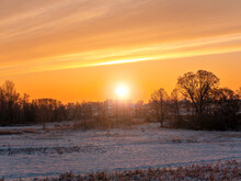 Sunny Sunset Over A Snowy Agricultural Field In Winter. Sunrise. White Snow. Cloudy Horizon. Sun Rays. Natural Winter Landscape. Agriculture. Background Image. Trees On The Horizon. Frosty Meadow.