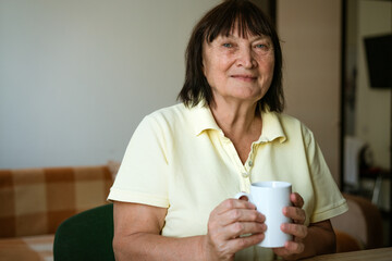 Mature woman sitting at table at home with mug in kitchen