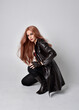 full length portrait of girl with long red hair wearing dark leather coat, corset and boots. Sitting pose facing front on with  magical hand gestures against a studio background.
