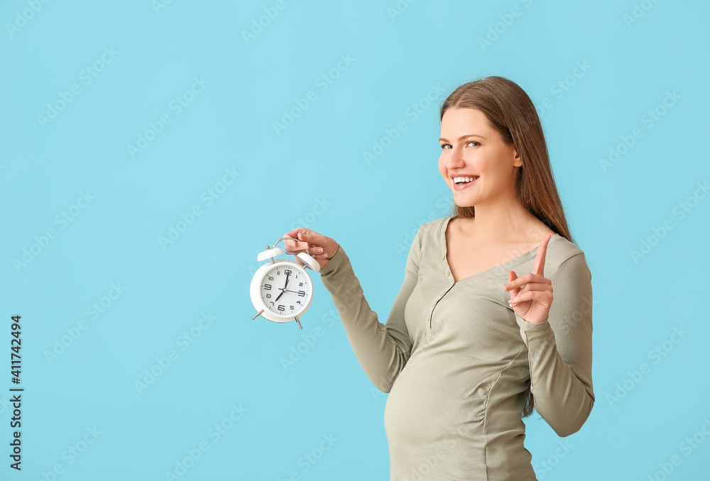 Pregnant young woman with clock on color background