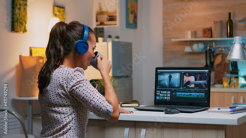 Woman video editor with headset working with footage and sound sitting in home kitchen. Woman videographer editing audio film montage on professional laptop sitting on desk in midnight © DC Studio