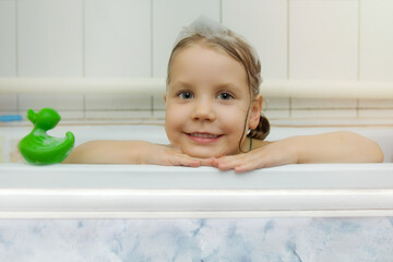 Children's hygiene. A beautiful little girl with foam on her head and a green duck for bathing looks out of the bath. The child bathes and smiles sweetly.