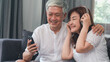 © tirachard - Asian senior couple relax at home. Asian Senior Chinese grandparents, husband and wife happy wear headphone using mobile phone listen to music while lying on sofa in living room at home concept.