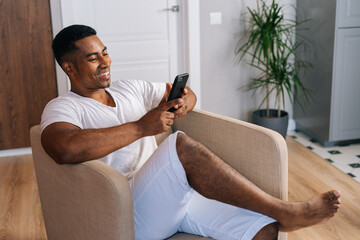 Poster - Close-up side view of cheerful African-American man in white T-shirt sitting at armchair in cozy bright domestic room and using mobile phone. Handsome male browsing internet on cellphone.