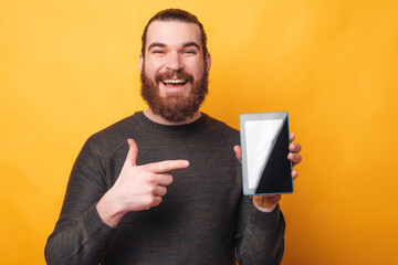 Photo of joyful man with beard pointing at tablet over yellow background.