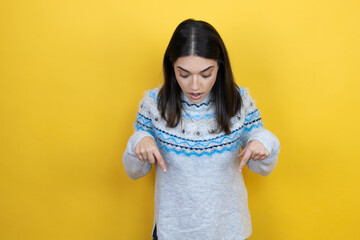 Wall Mural - Young caucasian woman wearing casual sweater over yellow background surprised, looking down and pointing down with fingers and raised arms