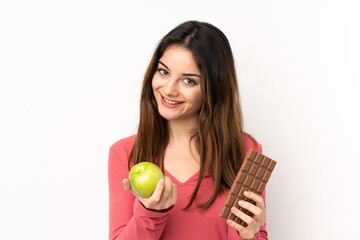 Canvas Print - Young caucasian woman isolated on pink background taking a chocolate tablet in one hand and an apple in the other