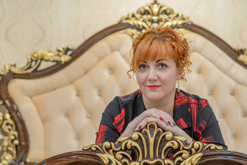 Poster - Beautiful red-haired woman lies on the edge of the bed with her arms folded on a wooden headboard an