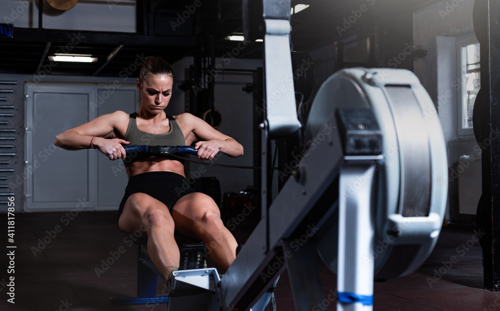 Young strong fit sweaty powerful attractive muscular woman with big muscles doing hard core row heavy training workout on indoor rower at the gym real people rowing exercise - obrazy, fototapety, plakaty 