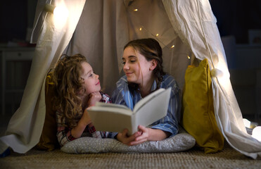 Wall Mural - Sisters indoors at home, reading book in pajamas. Lockdown concept.