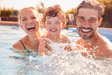 Portrait Of Family With Young Son Having Fun On Summer Vacation Splashing In Outdoor Swimming Pool