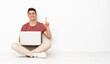© luismolinero - Teenager man sitting on the flor with his laptop showing and lifting a finger in sign of the best