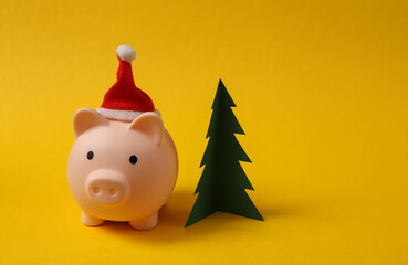 Wall Mural - Piggy bank with santa hat and Christmas tree on yellow background. Christmas concept