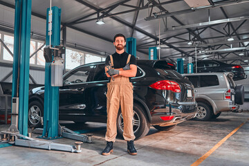 A professional male mechanical employee of a car service workshop stand with crossed arms and tools, looks very symphaticly and competently.