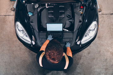 High angle view of mechanic examining car engine with help of laptop.