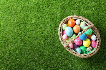 Wicker basket with Easter eggs on green grass, top view. Space for text