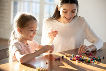 Happy Caring Young Caucasian Mother And Little 6s Daughter Have Fun Together Making Bracelets String Colorful Beads On Thread. Smiling Mom And Small Girl Child Involved In Funny Hobby Activity.