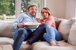 © Monkey Business - Couple On Date Night Sitting On Sofa At Home Watching Scary Movie On TV With Popcorn