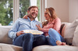 © Monkey Business - Couple On Date Night Sitting On Sofa At Home Laughing And Watching TV With Popcorn