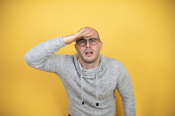 Young bald man wearing glasses over yellow background very happy and smiling looking far away with hand over head. searching concept.