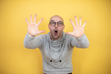 Wall Mural - Young bald man wearing glasses over yellow background showing and pointing up with fingers number ten while smiling confident and happy