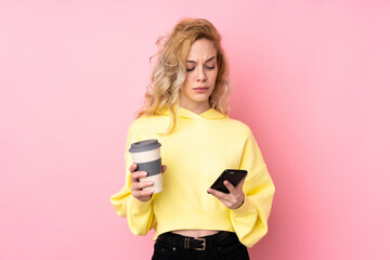 Poster - Young blonde woman wearing a sweatshirt isolated on pink background holding coffee to take away and a mobile