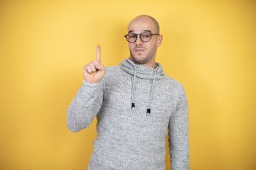 Wall Mural - Young bald man wearing glasses over yellow background showing and pointing up with fingers number one while smiling confident and happy