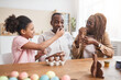 © Seventyfour - Portrait of loving African-American family making chocolate Easter decorations and tasting them while sitting at wooden table in cozy home interior