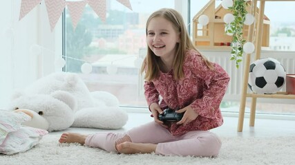 Poster - Smiling little girl playing video game with joystick sitting on floor in bright room