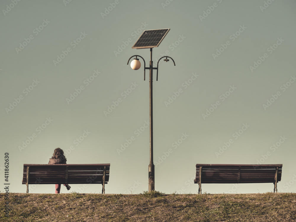Lonely woman sitting on a bench next to a light pole. Two benches with a solar panel lamp post in the middle - obrazy, fototapety, plakaty 