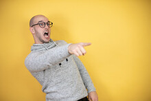 Young Bald Man Wearing Glasses Over Yellow Background Pointing With Finger Surprised Ahead, Open Mouth Amazed Expression, Something On The Front