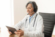 © Vadim Pastuh - Senior old female doctor in a lab coat sitting at the desk in her office with headset, consulting a patient on the insurance and treatment plan on a video call, telehealth or telemedicine concept