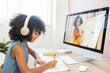 A clever multiethnic girl sits at table with a headphones, studying on the distance, doing homework, writes on workbook, watching online classes on the PC, side view. E-study, remote education