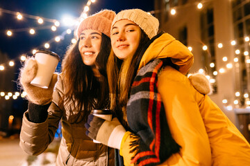 Two hipster female student hugging, drinking hot cocoa in winter with garland light around in night city street. Love, relationship, season, friendship and people concept.