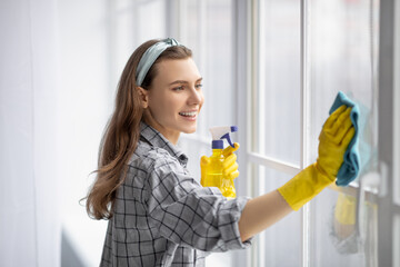 Portrait of young pretty lady in rubber gloves cleaning window with detergent at home