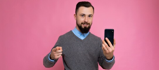 Panoramic photo of Handsome happy cool young brunette unshaven man with beardwearing stylish grey sweater and blue shirt standing isolated over pink background wall holding smartphone and using phone