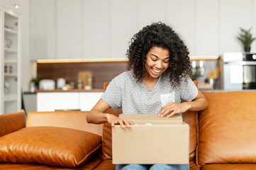 Positive young African American woman sitting on the couch with carton box on laps, feeling curious about ordered item from an online store, smiling client satisfied with fast courier delivery service