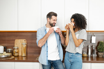 Shy young African American woman and handsome man interracial couple enjoying the happy morning together, standing leaning on the modern kitchen counter and holding cup of coffee in their hands