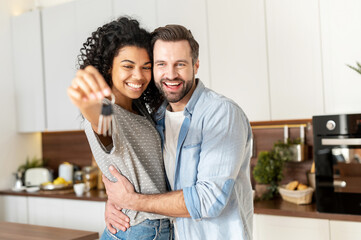 Young interracial married couple smiling cheerfully and showing keys from a new apartment, hugging and looking at camera, standing in the modern kitchen of a new home, real estate and family concept