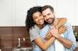 © Vadim Pastuh - Close-up of happy interracial couple posing over blurred kitchen background, happy owners of new flat smiling and looking at the camera, young African American woman hugging handsome man from behind